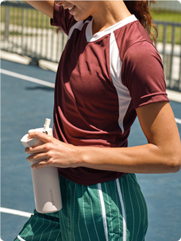 Woman in maroon athletic shirt holding a white Stanley 1913 water bottle on a tennis court
