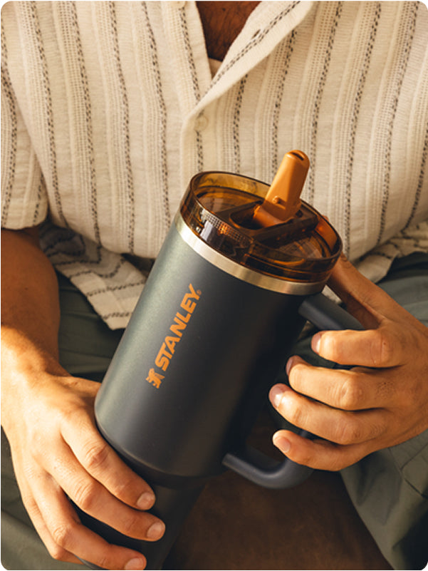 Stanley 1913 charcoal tumbler with amber lid and orange straw, held by a person in casual clothing.