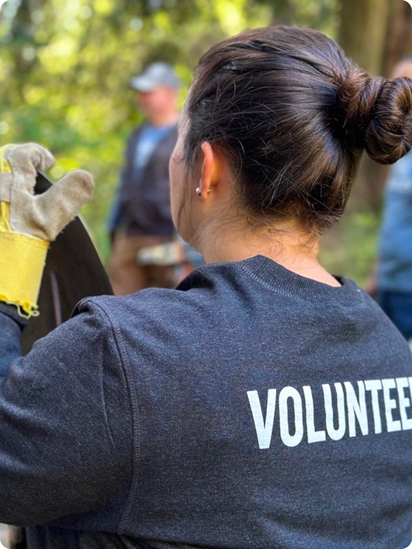 A volunteer in a dark gray "VOLUNTEER" shirt holds work gloves outdoors during a community service event.