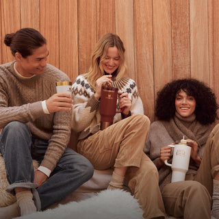 Three friends in cozy sweaters laughing together, each holding Stanley 1913 tumblers in cream, burgundy, and white.