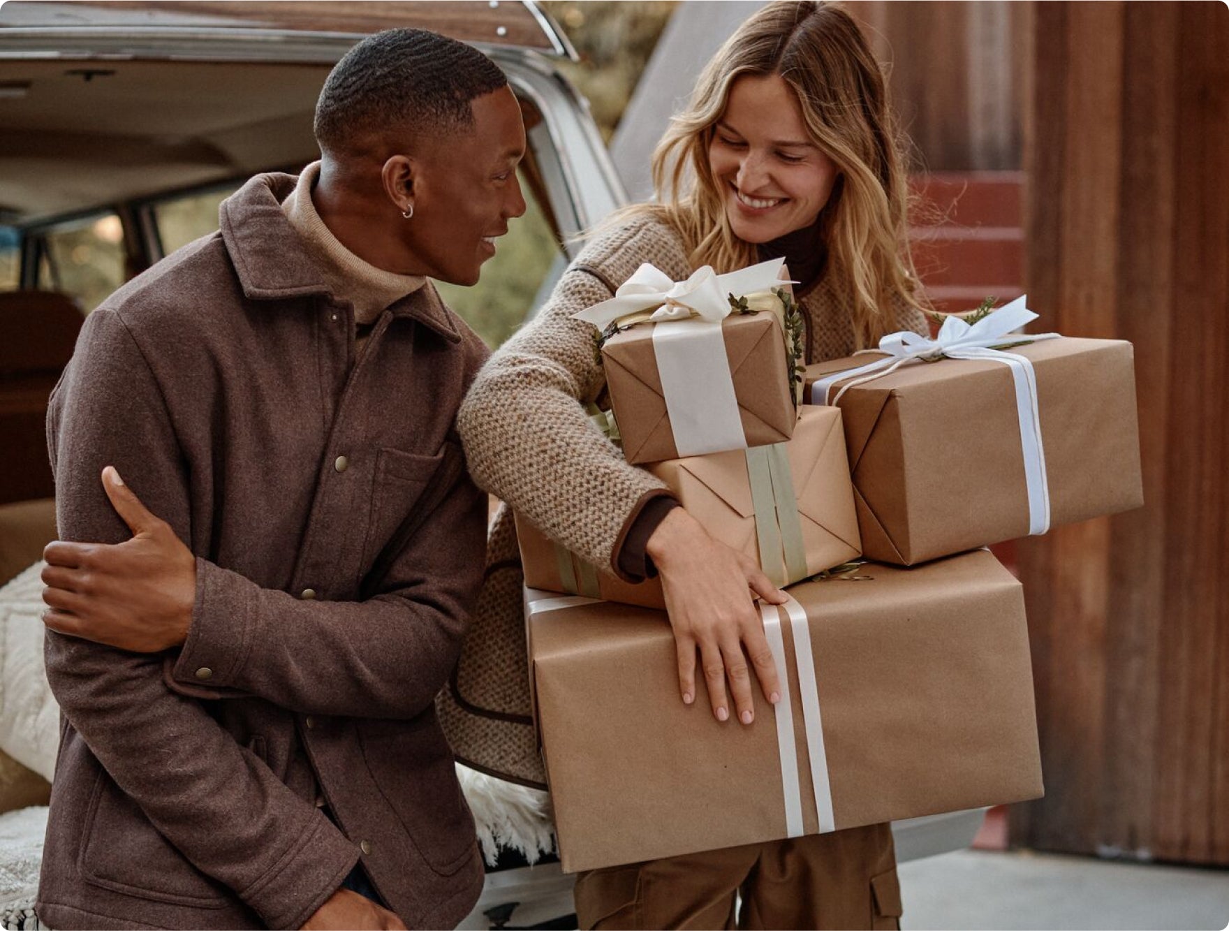 Two people carrying kraft paper-wrapped gifts with white ribbons, smiling together outdoors near a car trunk.