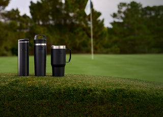 Stanley 1913 black insulated tumblers and travel mugs displayed on a golf course green with flag in background.
