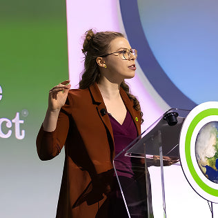 Young woman with glasses speaking at a podium at a professional conference, wearing a rust-colored blazer.