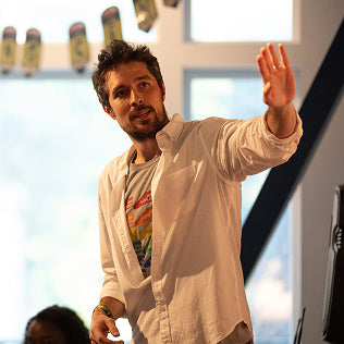 Man in white shirt gesturing expressively in a bright, energetic indoor setting.