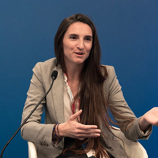 Woman speaking at a panel event, gesturing expressively, wearing a blazer, seated against a blue background.