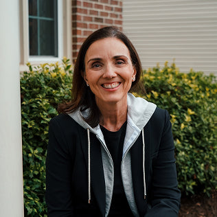 Woman smiling outdoors in a black zip-up hoodie, standing near greenery and a brick building.