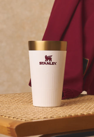 Stanley 1913 white stacking pint cup with gold rim and maroon logo, displayed on woven rattan surface with draped fabric.