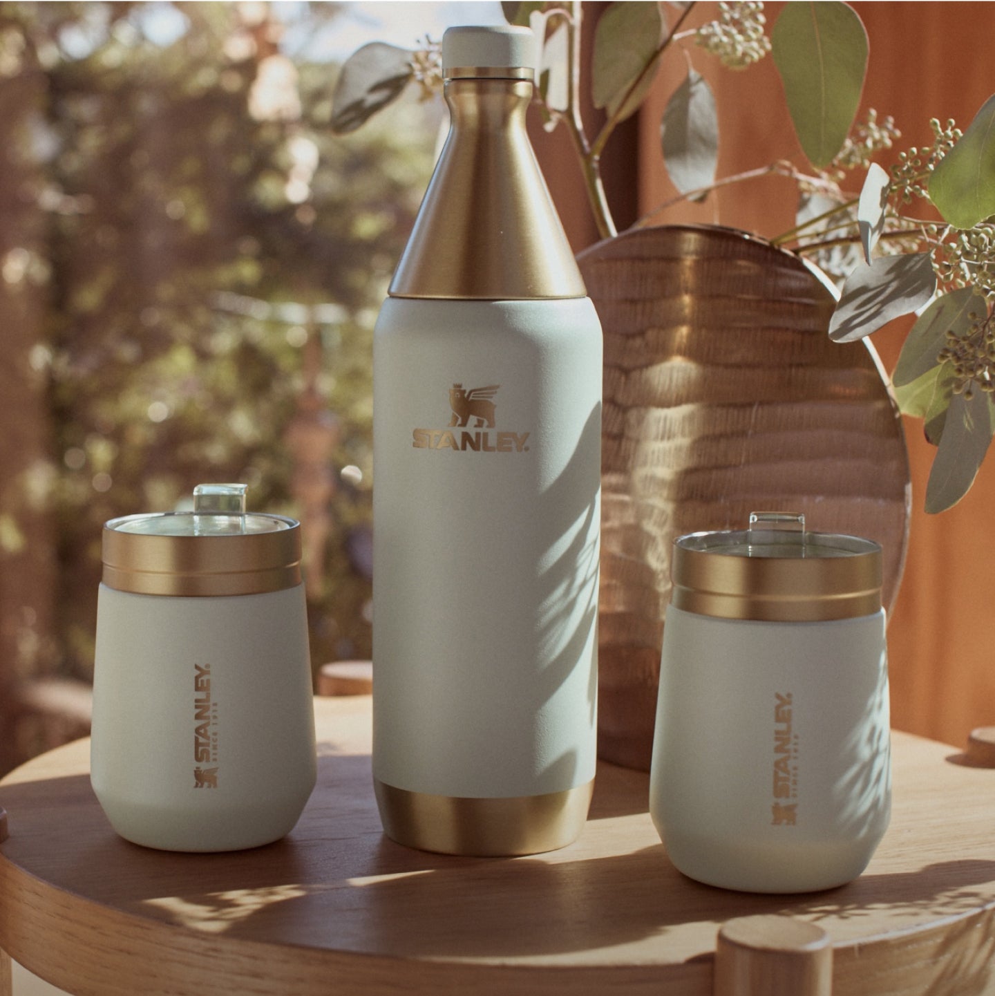 Stanley 1913 cream and gold insulated bottle with two matching tumblers on a wooden table in warm outdoor setting