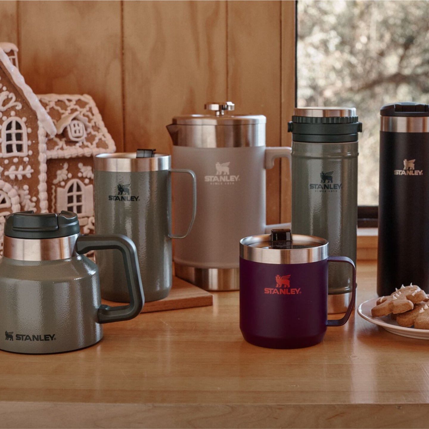 Stanley 1913 drinkware collection in slate, green, and plum displayed on a wood table beside a gingerbread house and cookies.