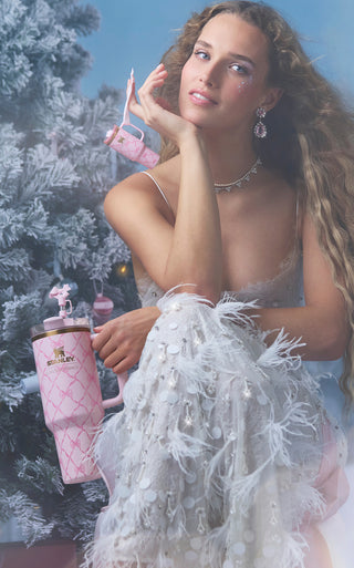 Stanley 1913 x Loveshackfancy pink bow-print tumbler held by a woman in a white feathered gown near a frosted Christmas tree