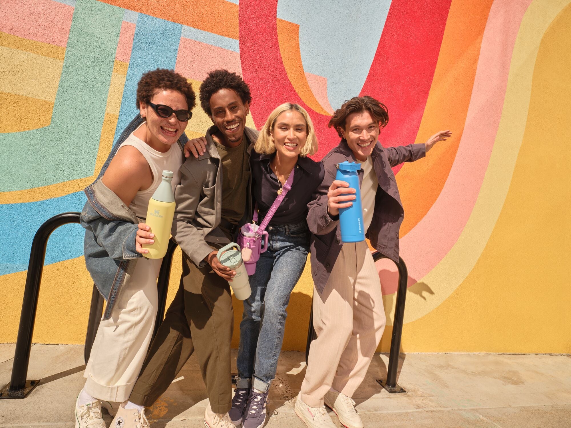 Four friends laughing in front of a colorful mural, each holding Stanley 1913 drinkware in pastel and bright colors.