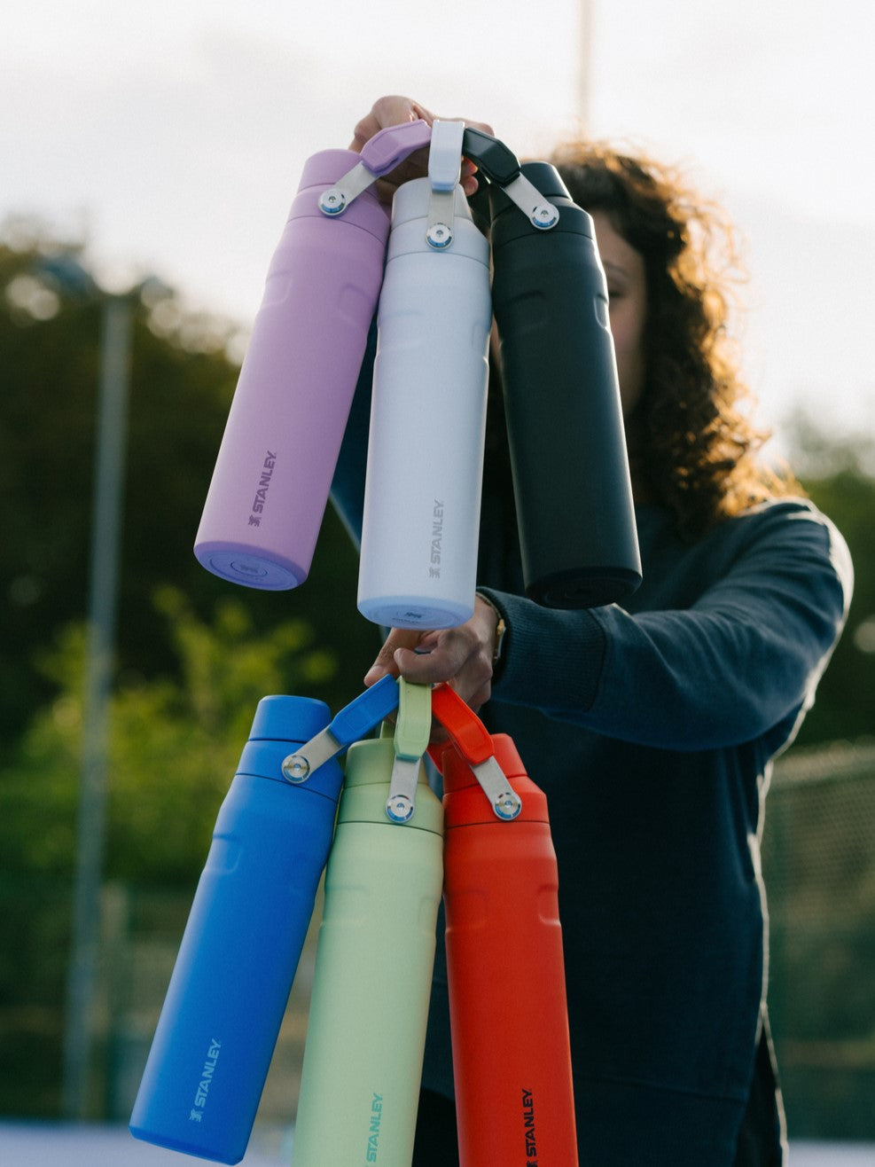 Person holding 6 Stanley 1913 water bottles in purple, white, black, blue, mint, and red with loop handles outdoors